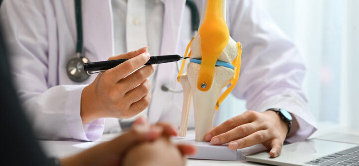 Orthopedists pointing at knee joint model, explaining the problem to patient during appointment.